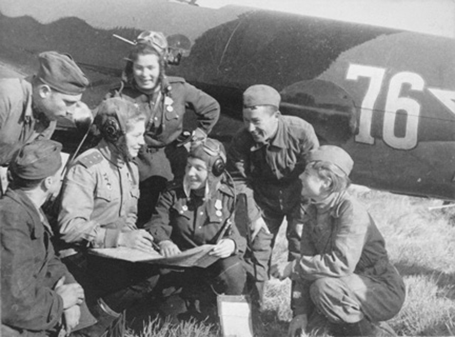 Members of a WWII bomber crew huddled together view mission documents in front of a bomber